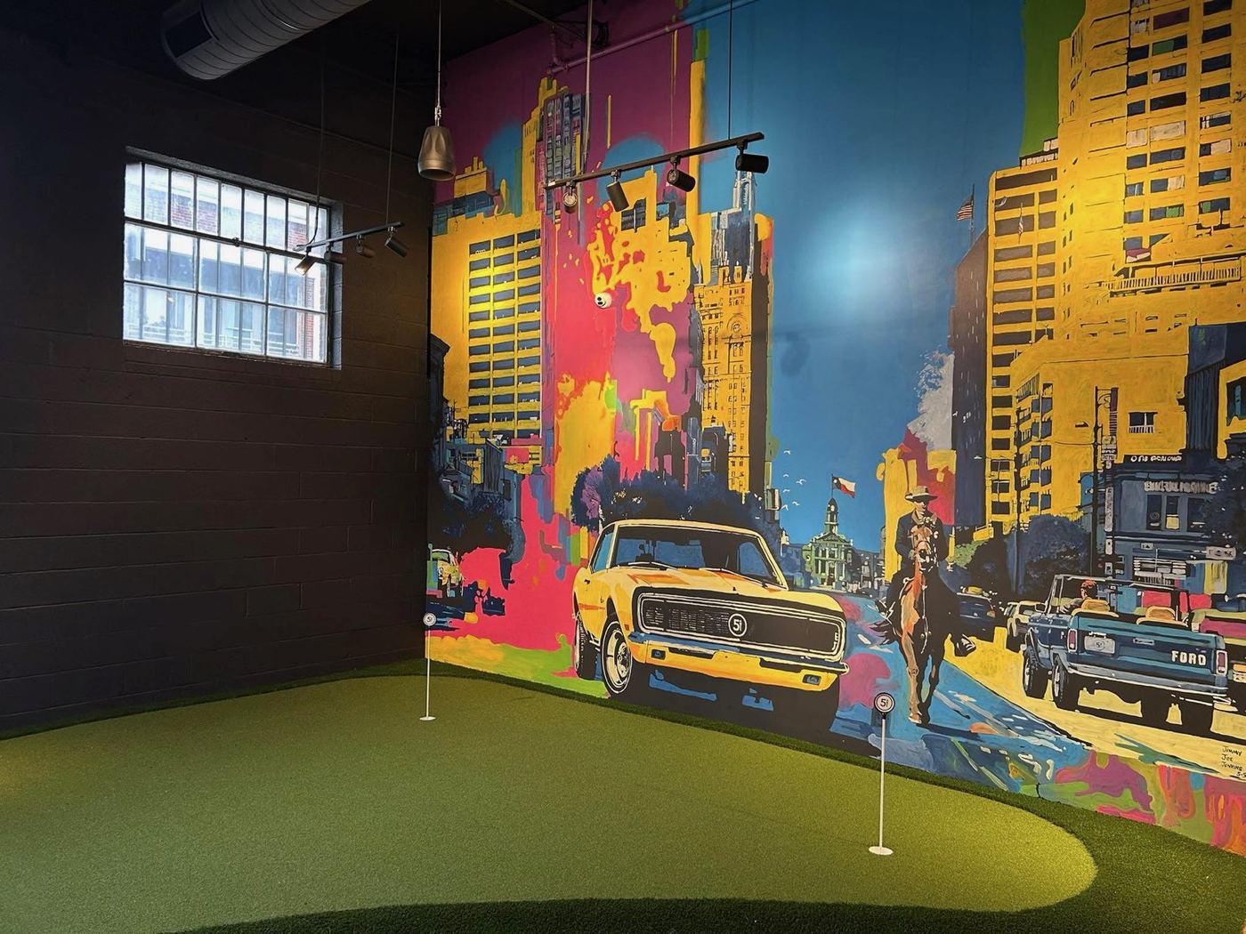 Indoor putting green at Five Iron Golf Fort Worth, backed by a mural of downtown Fort Worth with a cowboy on horseback and a yellow pickup truck.